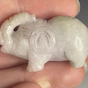 May include: A carved, off-white figurine of a small animal, possibly a pig or elephant. The figurine has rounded features, with etched details suggesting eyes, ears, and a circular design on its side. The sculpture is held in a person's hand.