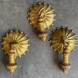 May include: Three antique gold-colored decorative wall sconces. Each sconce features a sunburst design with a central decorative element and a hanging pendant. The sconces are made of a metallic material and have a distressed finish.