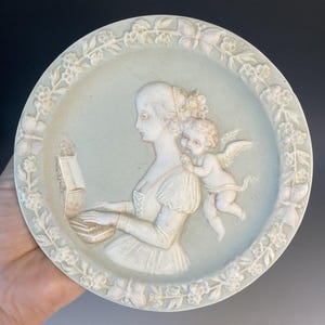 May include: A light blue ceramic plate with a raised relief design. The design features a woman playing a keyboard instrument, with a cherub on her shoulder. The plate has a floral border and is approximately 8 inches in diameter.