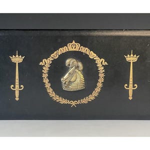 May include: A black rectangular box with gold embellishments. The center features a gold emblem of a knight's helmet within a laurel wreath. Two gold swords with crowns flank the emblem, creating a regal design.
