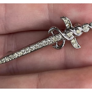 May include: Silver-toned brooch in the shape of a sword, encrusted with clear rhinestones. The brooch features a decorative hilt with curved accents and a series of rhinestones along the blade. A classic accessory.