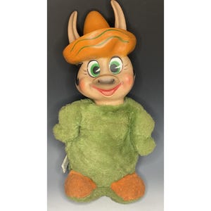 Puede incluir: Un juguete de peluche con un cuerpo de felpa verde y pies de fieltro naranja. El juguete tiene una cara pintada con una nariz marrón, ojos verdes y una amplia sonrisa. El juguete lleva un gran sombrero de fieltro naranja.