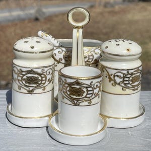 Może przedstawiać: Zestaw solniczki i pieprzniczki z białej porcelany z pozłacanymi zdobieniami i kwiatowym wzorem. Zestaw zawiera małą miseczkę do maczania. Solniczki mają mały otwór na górze do dozowania soli i pieprzu.