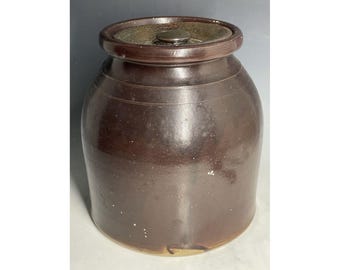 Puede incluir: Un tarro de barro marrón con tapa. El tarro tiene una forma ligeramente redondeada y una superficie lisa y vidriada. La tapa es de metal y tiene un simple pomo.