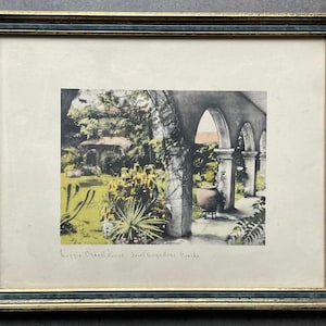 May include: A framed print depicting a garden scene with stone arches and a large clay pot, featuring yellow and green foliage. The text 'Oldest House Saint Augustine Florida' is written in pencil at the bottom of the off-white matting.