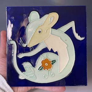 May include: Square ceramic tile with a stylized mouse design. The mouse is light green, cream, and beige, set against a deep blue background. A small orange flower with green leaves adds a pop of color.