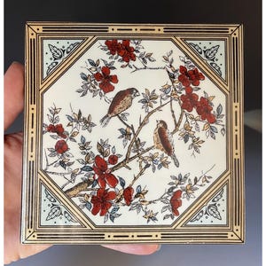 May include: A square ceramic tile featuring a cream-colored center with a detailed illustration of two brown birds perched on branches with red flowers and green leaves. The tile is framed by a geometric border in black, gold, and light blue.