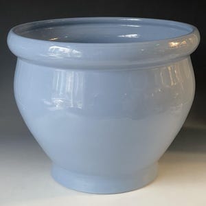 Puede incluir: Una maceta grande de cerámica azul claro, con un cuerpo redondeado y un borde ancho ligeramente ensanchado. La maceta tiene un acabado liso y brillante y una base robusta, adecuada para uso en interiores o exteriores. La forma general recuerda a una urna clásica.