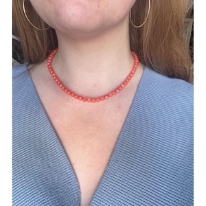 May include: A coral-colored beaded necklace. The necklace is composed of small, round beads and rests around the neck. The person is wearing a light blue sweater and gold hoop earrings.