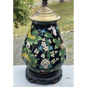 Op de afbeelding: Een decoratieve tafellamp met een zwarte voet en een messingkleurige bovenkant. De lamp heeft een zwarte behuizing met een bloemenontwerp in geel, roze en blauw. De lamp is ongeveer 46 cm hoog.