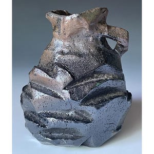May include: A handcrafted ceramic vase with a textured, faceted design. The vase features a dark, metallic finish with shades of black, gray, and brown. The unique shape and surface texture create a striking, modern aesthetic. A decorative piece for the home.