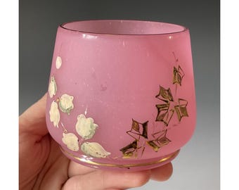 May include: Pink glass tumbler with a gold rim and floral design. The tumbler features white and gold painted flowers and leaves. The glass has a frosted appearance.