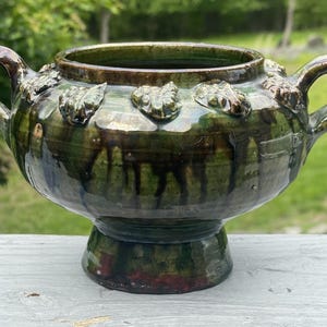 Peut inclure: Un bol en céramique émaillée avec deux anses et un socle. Le bol est principalement vert avec des accents marron et rouges. Des ornements décoratifs en forme de feuilles ornent le bord. Le bol est posé sur une surface blanche avec un fond vert.