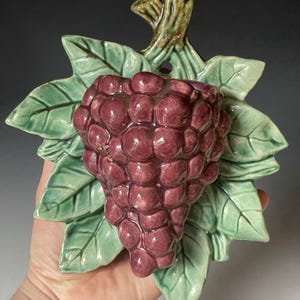 May include: A ceramic wall pocket shaped like a cluster of deep purple grapes surrounded by light green leaves. The piece features a glossy finish, a sculpted stem at the top, and a small circular hole for hanging on a wall or vertical surface.