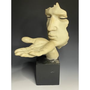 Puede incluir: Una escultura de color crema de un rostro humano y una mano. El rostro está parcialmente esculpido, con los ojos cerrados y una expresión serena. La mano está abierta, como ofreciendo algo. La escultura está montada sobre una base cúbica negra.