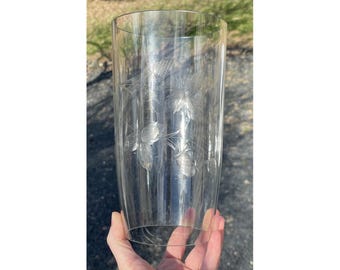 May include: A clear glass vase with an etched floral design. The vase is cylindrical in shape and has a smooth, polished finish.