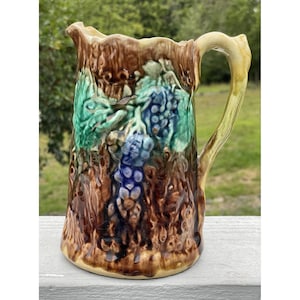 May include: A brown, green and blue ceramic jug with a grapevine design. The jug has a textured surface and a handle that is shaped like a branch.