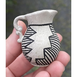 May include: A small, off-white ceramic jug with a handle and spout. The jug is decorated with black geometric patterns, including zigzags and stripes. The handle has a decorative, curved design.