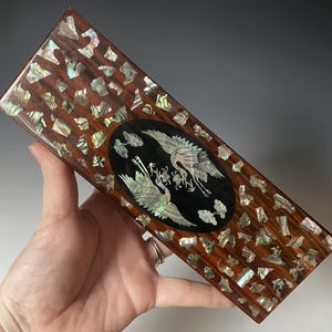 May include: Rectangular wooden box with a dark brown finish, inlaid with iridescent shell fragments. An oval centerpiece features two cranes in white and black. The box is held in a hand.