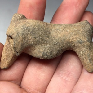 May include: A small, hand-carved stone sculpture of a stylised animal. The sculpture is made of a light brown stone with a rough texture. It is approximately 7.5 cm long and 5 cm wide.