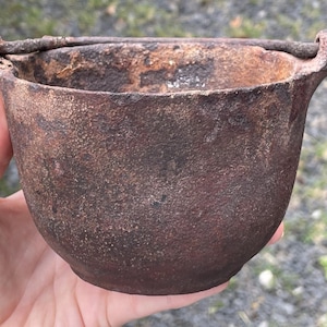 Peut inclure: Un petit chaudron antique en fonte avec une surface brun foncé et texturée. Le chaudron a un corps arrondi, un bord évasé et une poignée. La poignée est faite d'un fin morceau de métal incurvé. Le chaudron est tenu dans une main.