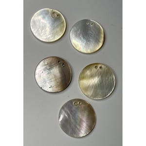 Könnte beinhalten: Fünf runde, schillernde Perlmutt-Anhänger mit zwei kleinen Löchern zum Auffädeln. Die Anhänger haben einen natürlichen, perlmuttartigen Glanz mit Farbvariationen, die von Weiß über Grau bis Braun reichen.