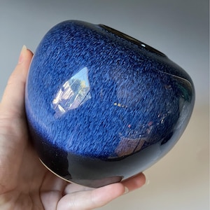 Puede incluir: Un jarrón de cerámica azul con acabado brillante. El jarrón tiene forma redondeada con una base azul oscuro que se desvanece a un azul más claro hacia la parte superior. La abertura es de color oscuro.
