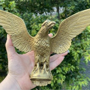 May include: A gold-colored metal eagle figurine with wings fully extended in a spread position. The eagle features detailed feather textures on its wings and body, with its head turned to the right and talons resting on a small, rounded base.