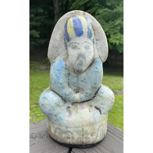 May include: A ceramic statue of a seated figure with a large headdress. The statue is painted in shades of blue, yellow, and white, with visible cracks and wear. The figure has a serene expression and is likely an ancient artifact.