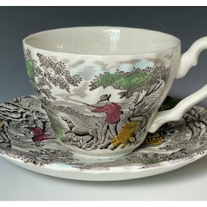 Può includere: Un set di tazza da tè e piattino in ceramica bianca con una stampa dettagliata in bianco e nero raffigurante un cacciatore con un cane in un paesaggio. La scena include accenti dipinti a mano in rosso, verde e giallo sul bordo smerlato.