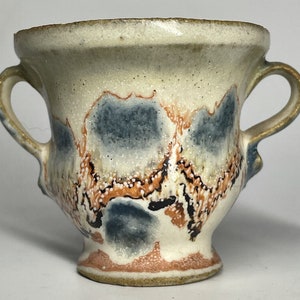 Può includere: Una piccola tazza in ceramica bianca con due manici. La tazza ha un design astratto unico con accenti blu, arancioni e neri.