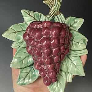 May include: A ceramic wall pocket shaped like a cluster of deep purple grapes surrounded by light green leaves. The piece features a glossy finish and a sculpted stem at the top, designed to be mounted on a wall for decorative storage or display.