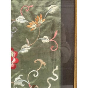 May include: A close-up of a green silk fabric with embroidered flowers in shades of pink, white, and orange. The fabric is framed in a dark wood frame.