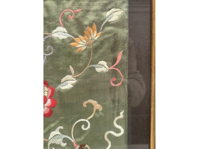 May include: A close-up of a green silk fabric with embroidered flowers in shades of pink, white, and orange. The fabric is framed in a dark wood frame.