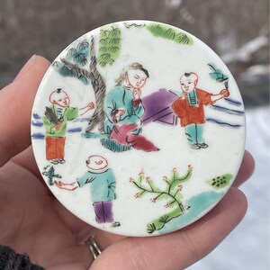 May include: A round porcelain plate with a white background and a colorful scene of four people in a garden setting. The people are painted in a traditional Chinese style and are wearing colorful clothing. The plate is decorated with green and blue trees and flowers.