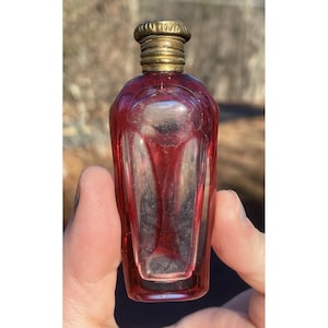 Puede incluir: Una pequeña botella de vidrio vintage de color rojo con una tapa de metal dorado. La botella tiene un diseño facetado y probablemente se usa para perfume o aceites esenciales.