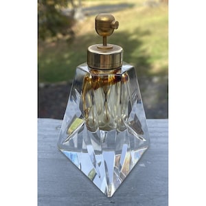 Peut inclure: Un flacon de parfum en verre clair et facetté avec un bouchon et un dessus en métal doré. Le flacon a un design géométrique et une base triangulaire. L'intérieur a une teinte dorée.