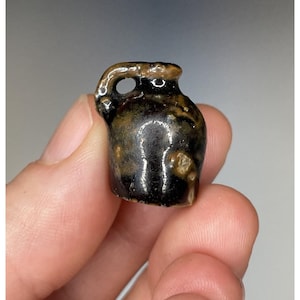 Puede incluir: Una jarra de cerámica en miniatura con asa y pico. La jarra es de color marrón oscuro con parches marrones claros y beige. La jarra está sostenida en la mano de una persona.