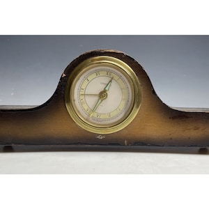 May include: A vintage wooden clock with a brass face and Roman numeral markings. The clock face is white with black markings and a blue hand. The clock is on a wooden base.
