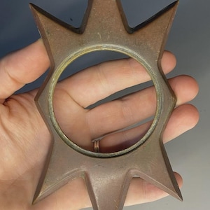 May include: A star-shaped metal object with six pointed rays extending from a central circular opening. The item features a weathered, brownish-bronze finish with visible signs of age and oxidation across its flat, angular surface.