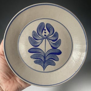 May include: A ceramic plate with a cream-colored base and a blue floral design in the center. The plate has a blue border and a hand-painted look. The plate is round and appears to be handmade.