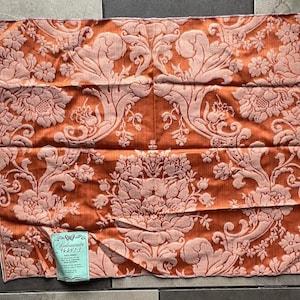 May include: A rectangular piece of fabric featuring a burnt orange background with a light pink damask floral pattern. A small, light blue rectangular tag is attached to the bottom left corner, displaying the text Salamandre 96247-3.
