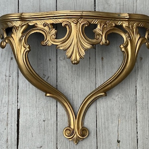 Könnte beinhalten: Verzierte goldfarbene Wandablage mit geschwungenem, herzförmigem Design. Das Regal weist aufwendige Schnörkel und ein dekoratives, blattartiges Mittelstück auf. Es ist auf einer verwitterten, hellfarbigen Holzoberfläche montiert.