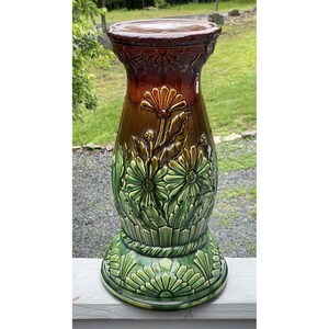 May include: A tall, ceramic plant stand with a detailed floral design. The stand features a dark brown top, transitioning to a lighter brown and then to green. The base and the floral details are in shades of green.