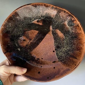 Puede incluir: Un plato redondo de cerámica con un esmalte brillante en tonos marrón oscuro, cobre y negro. La superficie presenta una textura moteada con patrones de pinceladas oscuras y pequeñas manchas negras dispersas a lo largo del borde.