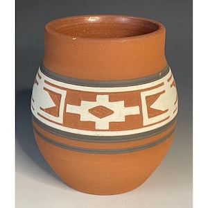May include: A terracotta-coloured ceramic vase with a wide mouth and rounded body. The vase features a geometric design in white and grey, with a band of repeating shapes around the middle. The design is reminiscent of Native American pottery.