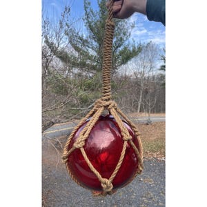 Pode incluir: Uma boia de vidro vermelha, grande e redonda, suspensa por uma rede de corda tecida e uma alça longa e grossa de corda. A boia é de um vermelho profundo e brilhante, e a corda é de cor bege natural. O fundo mostra árvores e um vislumbre de uma estrada.