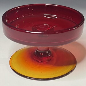 May include: A ruby red glass pedestal bowl with a wide, shallow top and a circular base. The base transitions from yellow to orange. The bowl is likely used for serving desserts or displaying decorative items.