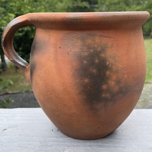 May include: A rustic, terracotta-colored pitcher with a rounded body and a curved handle. The surface has dark, mottled markings, adding to its aged appearance. The pitcher is likely handmade and suitable for decorative or functional use.
