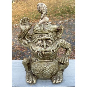 Puede incluir: Un caprichoso tarro de galletas de cerámica con forma de troll, con una mano levantada y una pequeña figura en la cabeza. El tarro es principalmente beige con texturas detalladas y una expresión sonriente. El tarro tiene una panza redonda con un patrón decorativo.